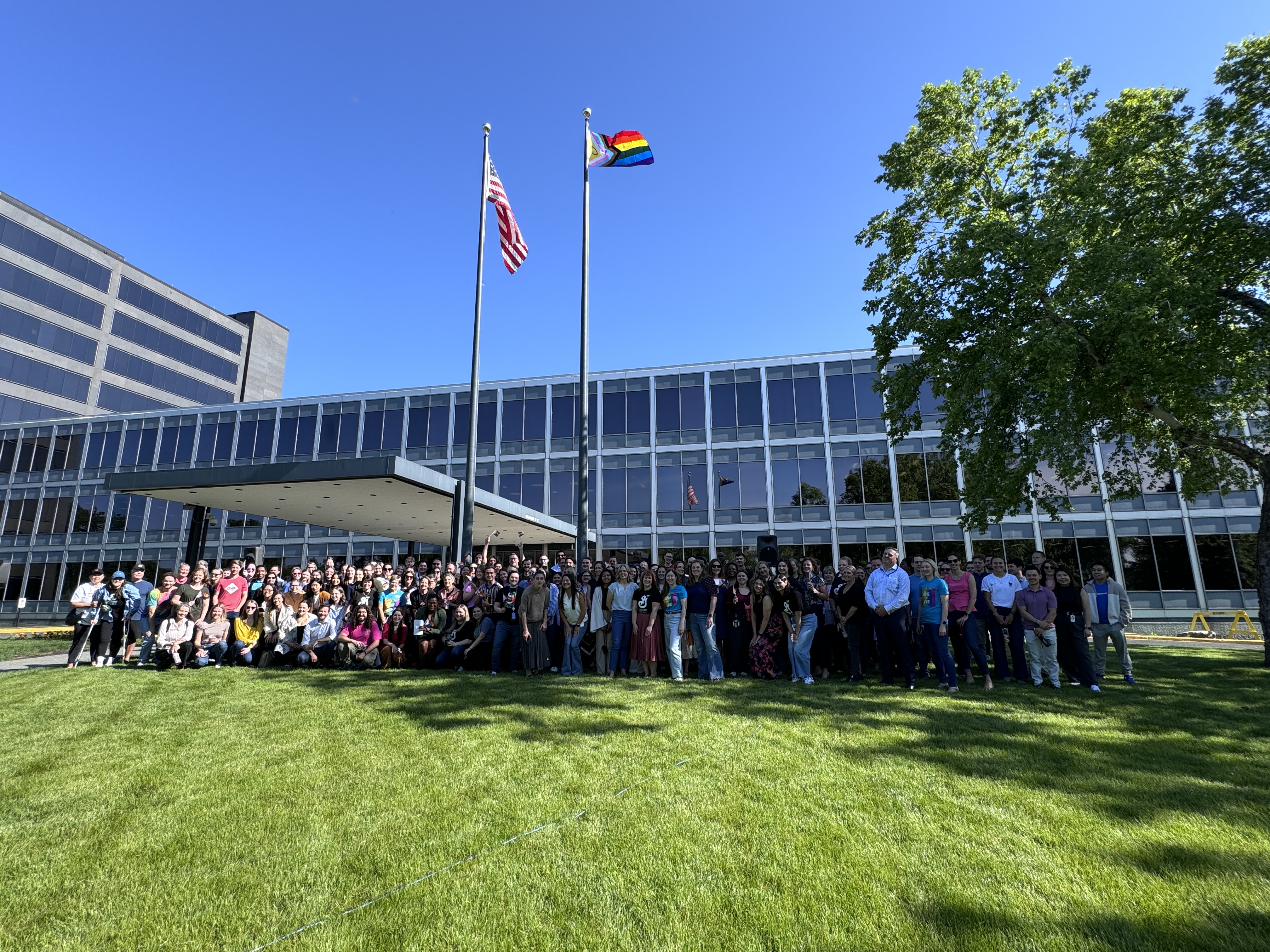 Group of General Mills employees gathering for a photo in front of the flag pole at corporate headquarters with the American and Pride flag flying proudly after Pride flag raising ceremony. 